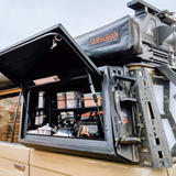 An Alu-Cab Kitchen Plate Assembled - Land Cruiser 76 mounted on the side window of a beige vehicle, with the black hardtop open to reveal cooking pots, containers, and a kettle inside.