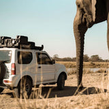 A white Jimny with Front Runner Suzuki Jimny (1998-2018) Slimline II Roof Rack Kit, is parked on a dirt road in a dry savanna landscape, with a large elephant standing close by in the foreground.