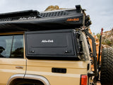 Beige vehicle with an Alu-Cab roof top tent, and the Alu-Cab Side Window Boxes to suit the Land Cruiser 76, in a rocky outdoor setting