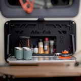 An open black Alu-Cab Alu-Cab INEOS Drop Down Table Mounting Kit mounted on a vehicle or equipment unit, holding several spice jars, containers, and two teal insulated tumblers on a metal shelf.