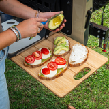 A wooden cutting board witch is part of the Alu-Cab INEOS Drop Down Table Mounting Kit shown outdoors on grass, holding prepared avocado toast slices with tomatoes and white cheese, alongside a person preparing more avocado on a sandwich. A grill is visible in the background.