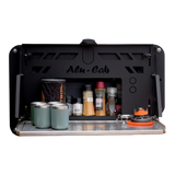 A black Alu-Cab INEOS Drop Down Table Mounting Kit shown open, with a metal tray shelf holding four teal tumblers, spice jars, and a small orange-and-black stove, on a white background.