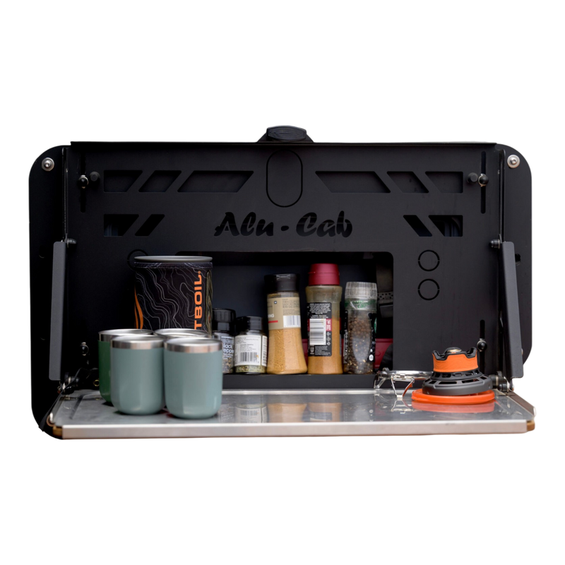 A black Alu-Cab INEOS Drop Down Table Mounting Kit shown open, with a metal tray shelf holding four teal tumblers, spice jars, and a small orange-and-black stove, on a white background.