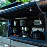An open Alu-Cab Kitchen Plate Assembled - Land Cruiser 76 reveals a compact outdoor kitchen setup with metal pots, containers, and cookware stored inside. Trees and outdoor scenery are visible through the vehicle windows.