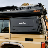 Black Alu-Cab rooftop storage box mounted on the side of a beige safari vehicle, with a black Alu-Cab roof rack above, photographed outdoors under an overcast sky.