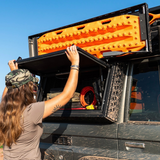 A woman in outdoor gear opens a Alu-Cab Universal Recovery Plate Assembled - Land Cruiser 76 on the side of a muddy off-road vehicle, with an orange recovery board mounted above on a roof rack against a blue sky.

