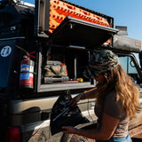A woman in a brown T-shirt is organizing gear inside an Alu-Cab Universal Recovery Plate Assembled - Land Cruiser 76 of a black off-road vehicle. The vehicle has a bright orange traction mat mounted on its roof rack, set against a background of blue sky and water.