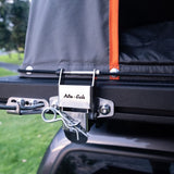 Close-up of a black Alu-Cab LT-50 Lightweight rooftop tent mounted on a vehicle, secured with a silver metal quick-release buckle labeled 'Alu-Cab' and an orange zipper trim, outdoors on grass.