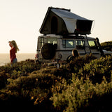 A white Camp4x4 rental off-road vehicle with an Alu-Cab RT-2S rooftop tent pitched above it, parked on a grassy hillside at sunset. Two people are visible in the outdoor setting, one standing and one seated near the vehicle.