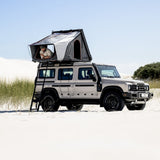 A beige Camp4x4 Rental Vehicle parked in a sandy desert setting with an Alu-Cab RT-2S rooftop tent up on the roof, and a person sitting inside the tent. The scene is bright and sunny under a clear blue sky.