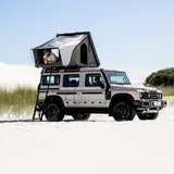 A beige Camp4x4 Rental Vehicle parked in a sandy desert setting with an Alu-Cab RT-2S rooftop tent up on the roof, and a person sitting inside the tent. The scene is bright and sunny under a clear blue sky.