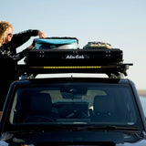 A Camp4x4 rental vehicle, fitted with the Alu-Cab LT-50 Lightweight rooftop tent, with a person securing surfboards to the roof