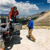 Three people unloading some beers from a Dometic CFX5 55 portable refrigerator from a truck in a mountainous area.