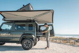Man setting up an Alu-Cab 270 Awning and Hard shell rooftop tent on a Jimny 4x4 by the beach