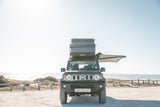Dark olive green Suzuki Jimny with a rooftop tent on a road near a beach.