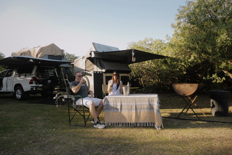 Two people sitting at a camp site outdoors drinking champagne from camping flutes and a bottle of champagne on ice.