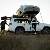A white SUV parked outdoors in a grassy field at sunset, with aFront Runner Feather-Lite II Rooftop Tent mounted on the roof rack and a ladder. A woman is climbing the ladder while another person stands by the open rear hatch.