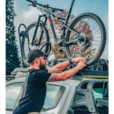 A man in a black t-shirt and cap is securing a black full-suspension mountain bike To a Front Runner Pro Bike Carrier, mounted on the roof of a silver pickup truck. The background shows trees and a cloudy sky.