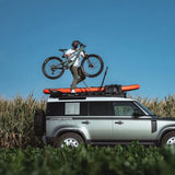 A silver SUV parked in a field under a clear blue sky, with a person standing on the roof rack carrying a mountain bike and holding an orange surfboard. The vehicle has roof-mounted gear and a Front Runner Pro Bike Carrier fitted