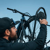 A man in a black cap and black long-sleeve shirt adjusts a black Front Runner Pro Bike Carrier with a bicycle with a Maxxis tire, set against a clear blue sky.