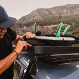 A man in a black t-shirt and navy cap is shown from the side, securing a surfboard with bright green and gray fins onto a Front Runner Pro Surfboard, Windsurf & Paddle Board Carrier on a  black roof rack of a silver off-road vehicle, with a mountainous landscape in the background.