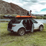 A white SUV parked on grass by a river with mountains in the background, its doors and trunk open, and a person sitting on an orange kayak mounted on a roof rack with the Front Runner Pro Canoe / Kayak / SUP Carrier.