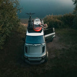 A silver Land Rover Defender parked outdoors by a river, with a person standing by an open side door and a bicycle and surfboard strapped to the roof rack.