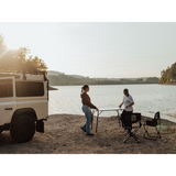 Two people setting up a Front Runner Under Rack Table, by a lake with a vehicle nearby.