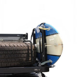 A close-up side view of a black roof rack on a vehicle, holding a blue and white surfboard with the Front Runner Vertical Surfboard Carrier, and a spare tire, against a plain white background.