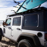 A white and black Jeep Wrangler parked on a sandy beach with a turquoise surfboard strapped to the Front Runner Vertical Surfboard Carrier on a roof rack .