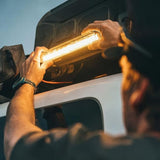A person installing a bright rectangular Front Runner Series 10" LED Light Bar VX250-FL / 12V/ 24V / Flood Light onto the side of a white vehicle at dusk, with the light glowing warmly against the dark vehicle body.