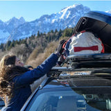 A woman in a blue jacket and black gloves loads a white and red hiking backpack into the Front Runner Cargo Carrier / 450, with a snowy mountain range in the background.