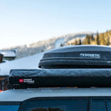 A black Dometic Front Runner roof top cargo carrier and matching soft cover mounted on the roof of a vehicle, shown outdoors with snowy mountains in the background.
