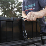 A person in a dark gray t-shirt secures a Front Runner Cub Pack Ammo Box with a black strap and silver carabiner in the bed of a truck outdoors.
