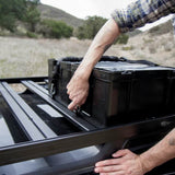 A Front Runner Cub Pack Ammo Box with a handle and straps being secured onto a black metal rack mounted on the bed of a truck, with a person's hands and arms visible in an outdoor desert landscape.