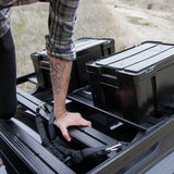 A person's arm resting on a black roof rack system carrying two Front Runner Cub Pack Ammo Box storage bins in the back of a vehicle, with an outdoor dry grass background.