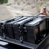 Two black Front Runner Cub Pack Ammo Box storage boxes strapped to the bed of a vehicle, with a hand holding a strap in an outdoor setting.