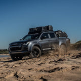 A dark gray Isuzu pickup truck drives off-road on sandy terrain, carrying a Front Runner Series Isuzu D-MAX RG/3rd Gen (2020-Current) Slimline II Roof Rack Kit loaded with black cases. The truck has 'Isuzu' branding on the front grille and 'Rover Accessories' decals on the side.