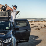 A man stands open-side up in a black 4x4 vehicle on a sandy beach, holding a large camera lens. The vehicle has a Front Runner Series Isuzu D-MAX RG/3rd Gen (2020-Current) Slimline II Roof Rack Kit and beach-themed decals, with the ocean and rocks visible in the background.