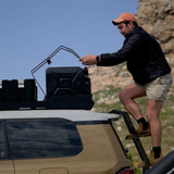 A man in outdoor gear stands on a ladder next to a vehicle, securing a black fuel can on a Front Runner Double Jerry Can Holder, mounted to a roof rack against a rocky, sunny mountain backdrop.