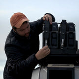 A man in a black puffer jacket and orange cap adjusts a black Front Runner Double Jerry Can Holder mounted on a vehicle, with a cloudy sky in the background.