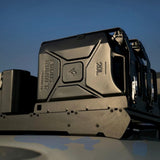 A close-up shot of a matte black Front Runner 20L container secured with a Front Runner Double Jerry Can Holder mounted on a vehicle, against a blue sky background.
