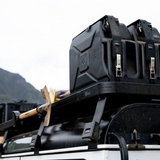 Black Front Runner Double Jerry Can Holder strapped to the roof rack of a vehicle, with an axe hanging from the side and a cloudy outdoor background.