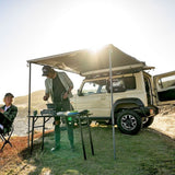 A lifestyle outdoor scene featuring a cream-colored off-road SUV with an extended canvas awning and a small folding table set up on a grassy hillside. Two people are present, one standing under the awning preparing food at the table and another sitting in a folding chair to the left.
