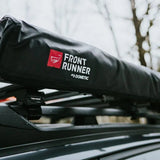 A black Dometic Front Runner awning mounted on a vehicle roof rack, with a blurred outdoor background of bare trees.