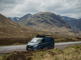 A dark blue Ford Transit camper van with a Front Runner Ford Tourneo/Transit Custom LWB (2013-2022) Slimline II Roof Rack Kit and roof-mounted camping setup drives along a road between rugged brown mountains under a cloudy sky.