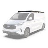 A white Ford Transit van shown at a three-quarter front angle on a light gray studio background, with a Front Runner Ford Transit Custom LWB (2023-Current) Slimsport Rack Kit / Lightbar Ready