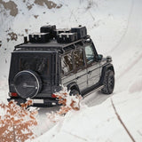 A black Mercedes-Benz G-Class SUV parked in deep snow, viewed from behind and above. The vehicle has a Front Runner Series Mercedes-Benz Gelandewagen G Class (1979-2017) Slimline II Roof Rack Kit loaded with black storage cases and a spare Mercedes-Benz tire mounted on the rear door.