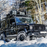 A black Mercedes-Benz G-Class SUV with a Front Runner Series Mercedes-Benz Gelandewagen G Class (1979-2017) Slimline II Roof Rack Kit and off-road tires driving through deep snow in a wooded winter setting.