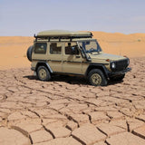 A beige off-road SUV with a Front Runner Series Mercedes-Benz Gelandewagen G Class (1979-2017) Slimline II Roof Rack Kit and a roof top tent is parked on cracked desert earth with sand dunes in the background.
